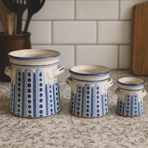 Könnte beinhalten: Drei unterschiedlich große Keramikvasen mit cremefarbenem Grund, blauen vertikalen Streifen und gepunkteten Akzenten. Jede Vase hat einen blauen Rand und kleine Griffe. Die Vasen stehen auf einer gesprenkelten Arbeitsplatte.