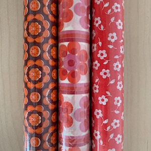 Könnte beinhalten: Drei Rollen Geschenkpapier nebeneinander. Die linke Rolle hat einen dunkelbraunen Hintergrund mit wiederkehrenden orangefarbenen und braunen Blumenmustern. Die mittlere Rolle hat einen weißen Hintergrund mit großen orangefarbenen und rosafarbenen Blumen. Die rechte Rolle ist rot mit kleinen weißen Blumen.