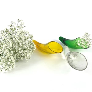 Könnte beinhalten: Drei glasförmige Vasen in Hornform in Gelb, Grün und Klar. Die grüne Vase enthält weiße Blumen. Ein großer Strauß weißer Blumen befindet sich links. Die Vasen stehen auf weißem Hintergrund.