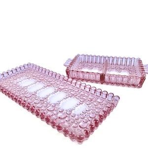 Könnte beinhalten: Zwei rosa Serviertabletts aus Glas. Eines ist rechteckig mit einem Blasendesign und weißen Ovalen. Das andere ist ein kleineres rechteckiges Tablett mit zwei Fächern und einem geriffelten Design. Auf weißem Hintergrund.