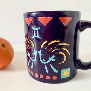 Könnte beinhalten: Eine dunkelviolette Keramiktasse mit Henkel, verziert mit farbenfrohen Tierkreiszeichen. Die Tasse hat rote Dreiecke, gelbe und blaue abstrakte Designs und einen Stern. Links von der Tasse liegt eine Orange.