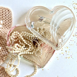 Könnte beinhalten: Eine transparente, herzförmige Glas-Schmuckschatulle mit Deckel, gefüllt mit Kunstperlen. Eine beige Spitzendecke und verstreute Konfetti sind ebenfalls im Bild. Die Schatulle befindet sich auf einer weißen Oberfläche.