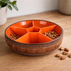 Puede incluir: Un cuenco redondo de cerámica naranja con un borde marrón estampado. El cuenco está dividido en seis secciones, llenas de almendras. Varias almendras están esparcidas sobre la superficie de madera.