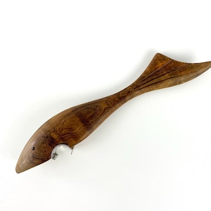 Könnte beinhalten: Ein Flaschenöffner aus Holz in Form eines Fisches. Der Öffner besteht aus dunkelbraunem Holz mit sichtbarer Maserung und einem silbernen Metallöffner. Der Fisch hat einen spitzen Kopf, einen gebogenen Körper und eine Schwanzflosse.