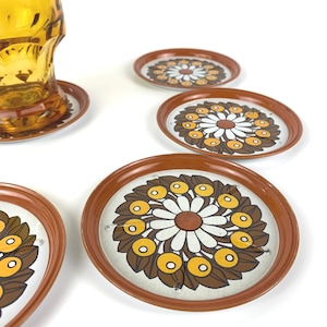 Könnte beinhalten: Sechs Vintage-Untersetzer mit einem braunen, weißen und gelben Gänseblümchen-Design. Die Untersetzer haben einen braunen Rand und liegen auf einer weißen Oberfläche. Ein bernsteinfarbenes Glas steht auf einem der Untersetzer.