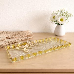 Könnte beinhalten: Ein rechteckiges Glastablett mit floralem Design und goldfarbenem Rand. Eine Perlenkette liegt auf dem Tablett, zusammen mit einem Spitzentuch und einer kleinen weißen Vase mit weißen Blumen.