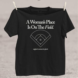 May include: Black t-shirt with the text "A Woman's Place Is On The Field" and a baseball field graphic. The phrase "support women in sports" is printed below the graphic. The shirt is hanging from a clothesline.