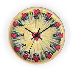 May include: Round wall clock with a light wood frame. The clock face features a burst of yellow and white light emanating from the center. Red hearts and the word "Now" are evenly spaced around the clock face.