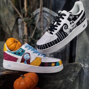 Puede incluir: Zapatillas blancas con ilustraciones personalizadas con personajes y temas de "Pesadilla antes de Navidad". Un zapato tiene a Jack Skellington, el otro a Sally. El texto "This is Halloween" es visible. Peque&ntilde;as calabazas en primer plano.