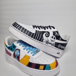 Puede incluir: Un par de zapatillas blancas con ilustraciones personalizadas. Una zapatilla presenta un dise&ntilde;o en blanco y negro con el texto "This is Halloween". La otra tiene un dise&ntilde;o de patchwork colorido y una ilustraci&oacute;n de personaje. Cordones azules.