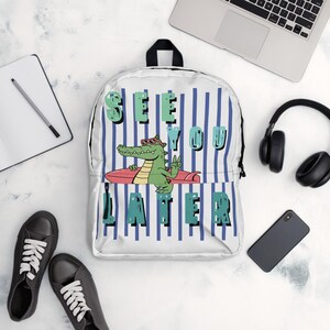 Puede incluir: Una mochila blanca con un patrón de rayas verticales azules y un gráfico de un cocodrilo verde con gafas de sol rojas sobre una tabla de surf rosa. El texto "SEE YOU LATER" está impreso en grandes letras verdes desgastadas al frente.