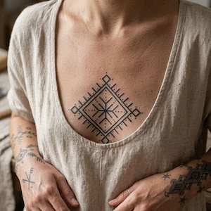Pode incluir: Uma pessoa usando um top de linho bege. Uma tatuagem geométrica preta em forma de diamante com um desenho de estrela está centrada no peito. Tatuagens adicionais são visíveis nos braços e mãos, juntamente com anéis de prata.