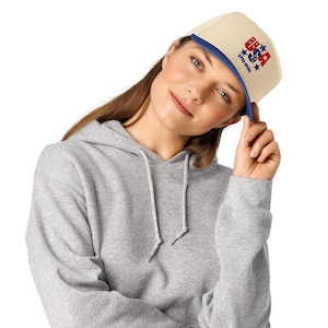 May include: A cream-colored flat-brimmed cap with a blue brim and the word "USA" in red and blue, along with stars and the years "1776-2026". The cap is worn with a gray hooded sweatshirt.