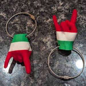 May include: Two hand-shaped keyrings. One keyring features a hand in the shape of the Italian flag, with red, white, and green colours. The other keyring is a hand gesture with the same colours. Both have a silver metal ring.