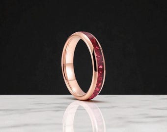 Könnte beinhalten: Roségoldener Ring mit einem Band aus leuchtend roten Steinen. Der Ring ist vor einem schwarzen Hintergrund platziert und spiegelt sich auf einer weißen Oberfläche. Das Design ist schlicht, betont aber die satte Farbe der Steine. Ein Schmuckstück.