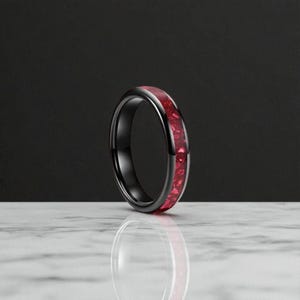 Peut inclure: Une bague noire avec une bande de pierres précieuses rouges. La bague est posée sur un fond sombre et se reflète sur une surface blanche. Il s'agit d'un bijou.