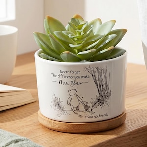 Pode incluir: Um vaso de cerâmica branco com uma planta suculenta verde, em uma base de madeira. O vaso tem uma ilustração de Winnie the Pooh e o texto "Never forget the difference you make Mrs. Glow". Uma caneca branca e um livro aberto estão por perto.