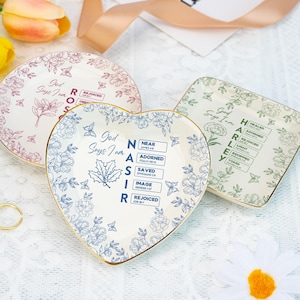 Puede incluir: Tres platos decorativos de cerámica de diferentes formas: redondo, corazón y cuadrado. Cada plato presenta motivos florales y ribetes dorados. El plato en forma de corazón tiene diseños florales azules y texto. El plato redondo tiene diseños florales rosas y texto.