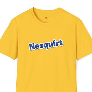 Peut inclure: T-shirt jaune moutarde avec le mot "Nesquirt" en bleu et blanc sur le devant. Le t-shirt a un col rond et un petit logo près de l'encolure.