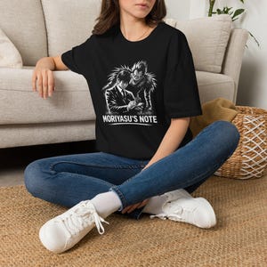 May include: A black short-sleeved t-shirt featuring a white graphic print of a person in a suit writing in a notebook next to a tall, spiky-haired creature. The text MORIYASU'S NOTE is printed in white capital letters below the graphic illustration.