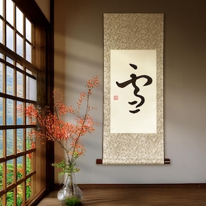 May include: A Japanese scroll with black calligraphy and a red seal hangs on a gray wall. The scroll is framed by a patterned border. A vase with red flowers and a window with a view of a green landscape are in the foreground.
