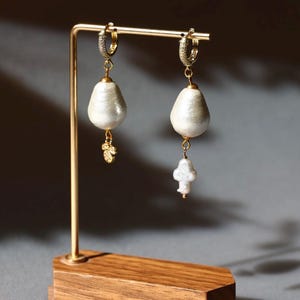 May include: A pair of elegant pearl earrings displayed on a gold-colored stand with a wooden base. Each earring features a large, teardrop-shaped pearl suspended from a small, diamond-studded hoop. One earring has a small gold charm.