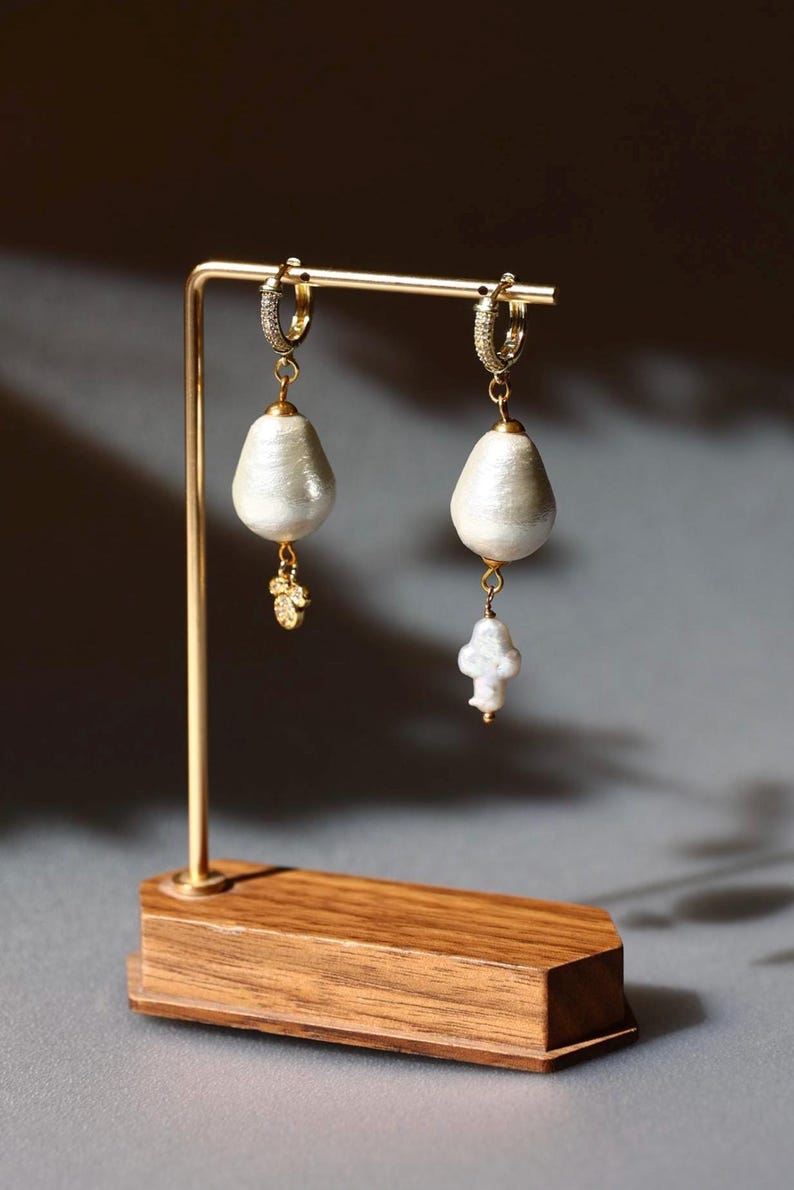 May include: A pair of elegant pearl earrings displayed on a gold-colored stand with a wooden base. Each earring features a large, teardrop-shaped pearl suspended from a small, diamond-studded hoop. One earring has a small gold charm.