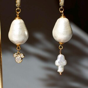 May include: Elegant gold-tone earrings with large, pearly white, teardrop-shaped pendants. One earring features a small, gold-tone charm with sparkling clear crystals. The other earring has a smaller, irregular pearl pendant.