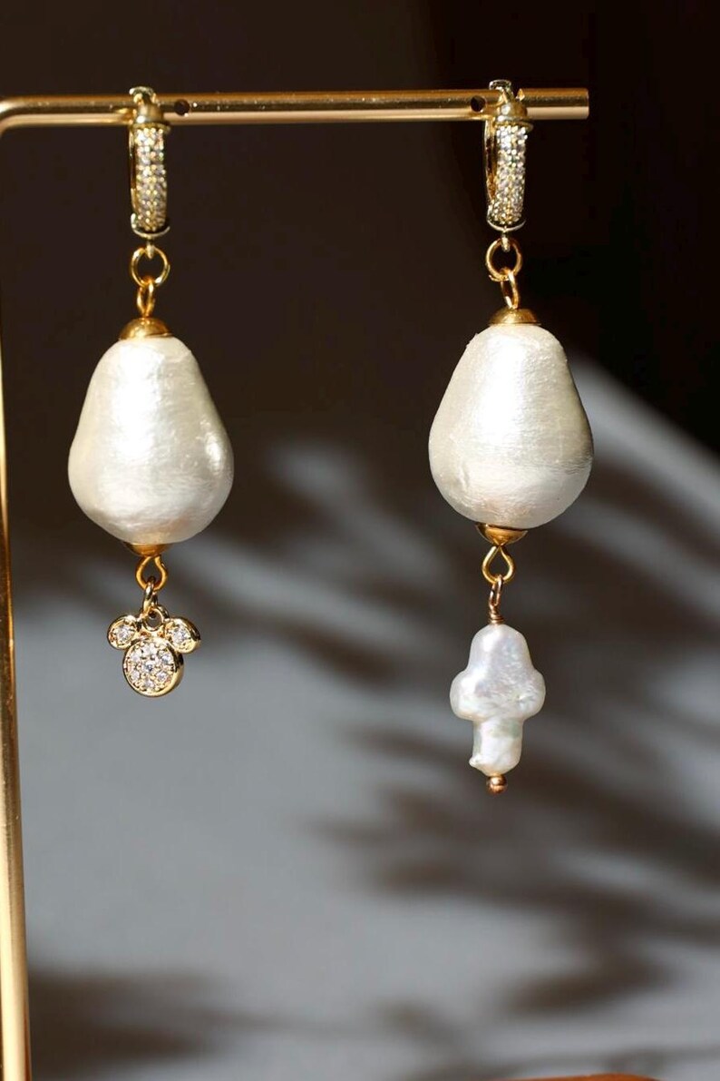 May include: Elegant gold-tone earrings with large, pearly white, teardrop-shaped pendants. One earring features a small, gold-tone charm with sparkling clear crystals. The other earring has a smaller, irregular pearl pendant.