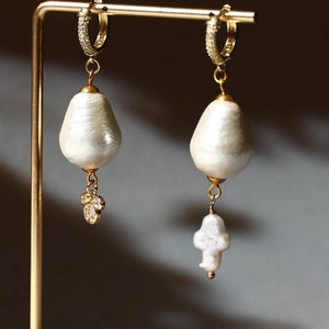 May include: A pair of gold-toned hoop earrings with pear-shaped, off-white pearl drops. Each earring features a small, sparkling charm and a larger, irregular pearl pendant, displayed on a gold stand.