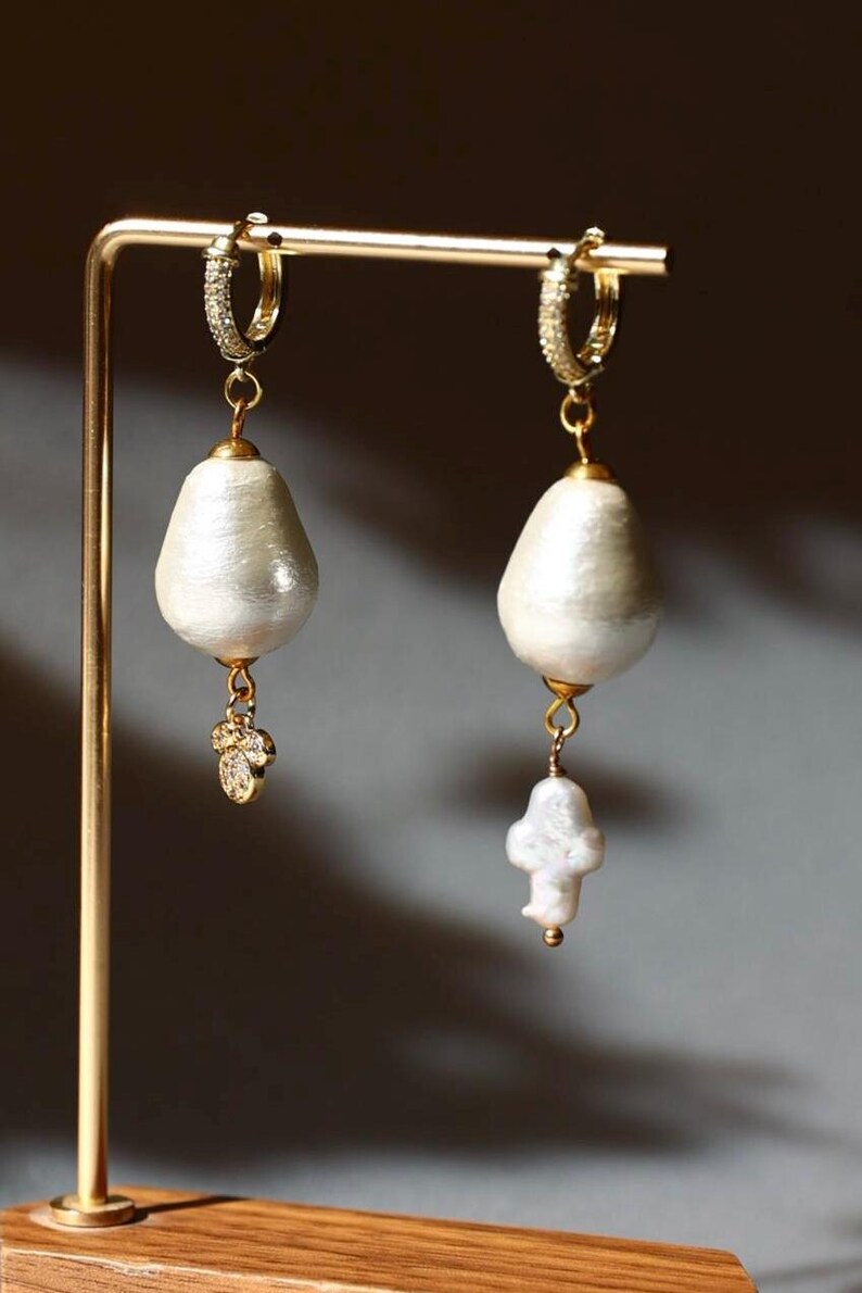 May include: A pair of gold-toned hoop earrings with pear-shaped, off-white pearl drops. Each earring features a small, sparkling charm and a larger, irregular pearl pendant, displayed on a gold stand.