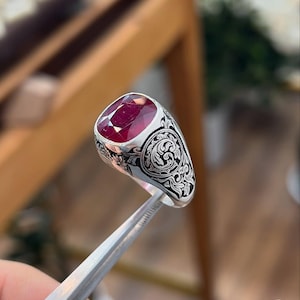 Puede incluir: Un anillo de plata con una piedra preciosa ovalada de color rojo intenso. El anillo presenta intrincados diseños de filigrana en negro y plata en la banda. El anillo está sujeto con unas pinzas, con un fondo borroso.