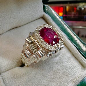 May include: A silver ring with a large, oval-cut ruby gemstone. The ruby is surrounded by a halo of smaller, clear gemstones. The ring has a detailed, textured band and is displayed in a green jewelry box.