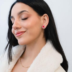 Puede incluir: Una mujer lleva pendientes cuadrados con un diseño de corazón rojo. Los pendientes tienen un fondo rosa. Lleva una chaqueta difusa de color crema y un collar de oro. Su pelo oscuro es largo y liso.