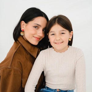 Puede incluir: Dos personas sonriendo a la cámara. Una persona lleva una chaqueta marrón y pendientes rojos y dorados. La otra persona lleva una camisa de manga larga de color claro y vaqueros azules, con pendientes dorados. El fondo es blanco.