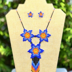 May include: A beaded necklace and matching earrings set. The necklace features three large blue flower pendants with orange centers. The necklace has a black beaded chain and a fringe of blue, orange, and black beads. The earrings are small blue flower studs.