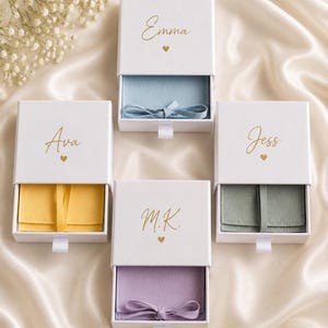 May include: Four white rectangular sliding jewelry boxes with gold foil names Emma, Ava, Jess, and M.K. printed on the lids above small gold hearts. Each box contains a fabric pouch in light blue, yellow, sage green, or lavender with a ribbon tie.