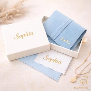 May include: A white gift box with the name "Sophia" in gold lettering. Inside, a light blue suede pouch and a cleaning cloth, also with "Sophia" in gold. A gold necklace with a pendant is visible.