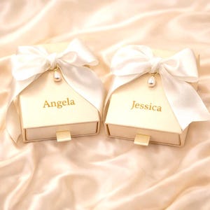 May include: Two ivory gift boxes, each with a white satin bow and a pearl charm. The boxes are personalised with the names "Angela" and "Jessica" in gold lettering. They have a small pull tab and are on a satin background.