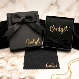 May include: Two black gift boxes with gold lettering that reads "Bridget". One box is closed with a black bow, the other is open. A black cleaning cloth and a gold necklace are also visible. The background is a marble surface with pearls.