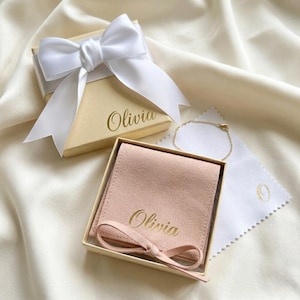 May include: A gift set featuring a small gold bracelet, a pale pink suede jewelry pouch, and a cream-colored box tied with a white satin bow. The box and pouch are inscribed with the name "Olivia" in gold lettering.