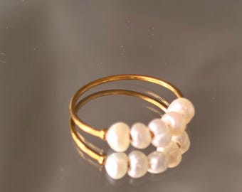 May include: A gold-colored ring featuring a row of six white and pink pearls. The ring's band is thin and circular, with the pearls evenly spaced across the top. The ring is displayed on a reflective surface.