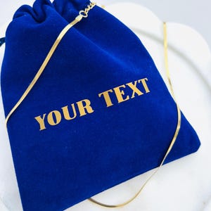 May include: A gold necklace lies across a royal blue velvet pouch. The pouch features the gold text "YOUR TEXT". The necklace is a thin, flat chain. The pouch is closed with a drawstring.