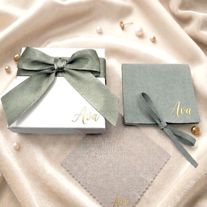 May include: A white gift box with a sage green ribbon and the name "Ava" in gold script. A matching sage green jewelry pouch and cleaning cloth are also present, each with "Ava" in gold script. The items are on a cream-colored satin surface.