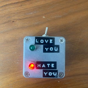May include: A small, silver box with a toggle switch on the top. The box has two red and green lights, and two black labels that read "LOVE YOU" and "HATE YOU". The red light is on.