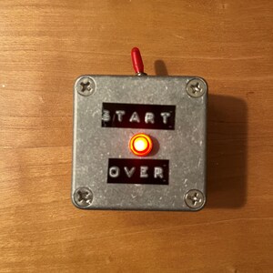May include: A small, square, metal device with the words "START" and "OVER" printed on it. A red light is illuminated in the center. A red lever extends from the top. The device is secured with four screws.