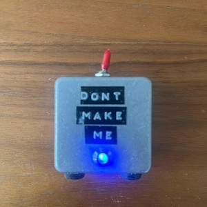 May include: A small, silver-colored box with a blue LED light. The box has the words "DON'T MAKE ME" printed on it.