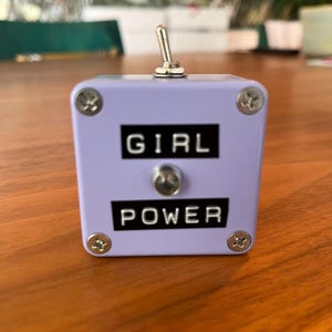 May include: A lavender-colored box with the words "GIRL POWER" in black lettering. The box has a silver toggle switch on top and is secured with four silver screws. The item is sitting on a wooden surface.