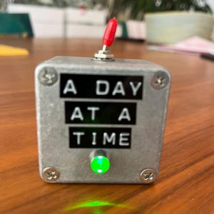 May include: A silver metal box with the words "A DAY AT A TIME" in black letters. It has a red toggle switch on top and a green light on the front. The box is secured with four screws.