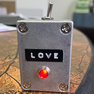 May include: A small, silver box with a toggle switch and a red LED light. The box has the word "LOVE" printed on it.